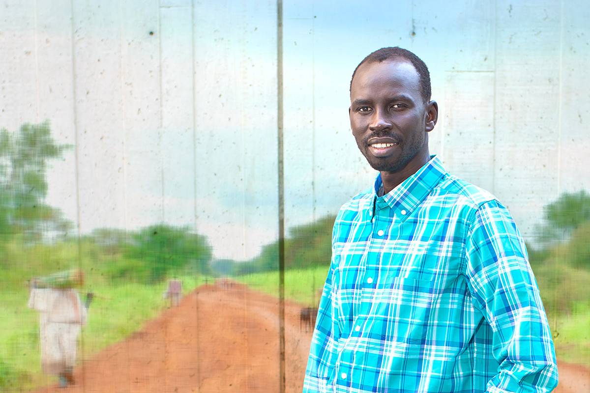 Garang Buk Buk Piol, stands in jeans and a light blue plaid button-down shirt in front of a mural depicting a red-dirt road in his native South Sudan, Africa.